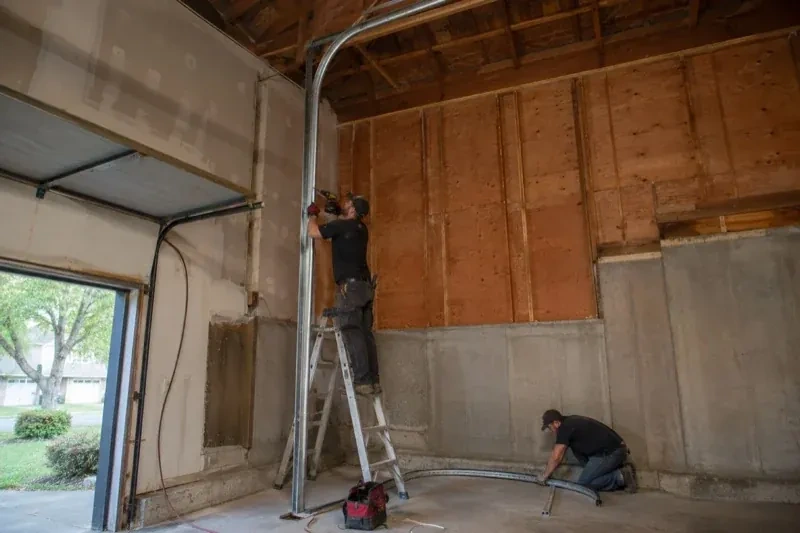 One person on a stepladder working beside a tall curved garage-door track while another kneels beside loose track sections in an unfinished garage. — High-Lift Garage Door Conversion, Port Colborne